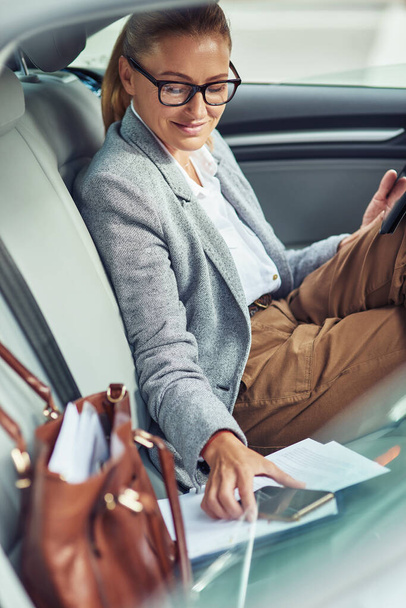 Foto vertical de una exitosa mujer de negocios sonriente con ropa clásica sentada en el asiento trasero del coche, usando su teléfono inteligente y sonriendo - Foto, Imagen