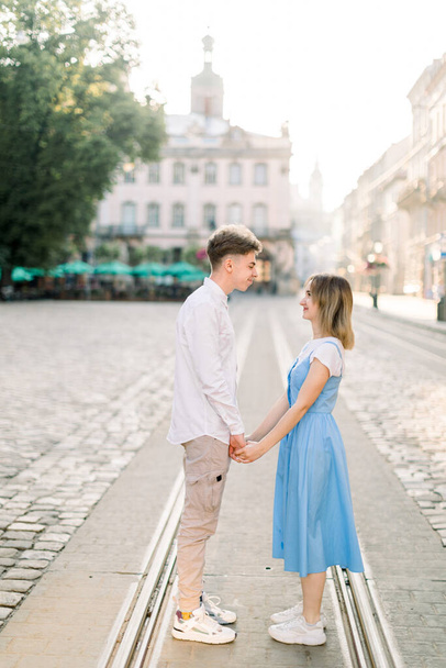Jsme rádi, že jsme spolu. Portrét krásného mladého páru, který se drží za ruce a dívá se na sebe s úsměvem, zatímco stojí na tramvajové dráze na dlážděné cestě ve starém evropském městě - Fotografie, Obrázek