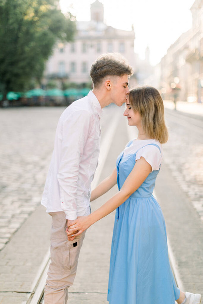 Handsome young man kissing his charming pretty girlfriend on forehead in the street in the sunny morning. Romantic couple in the street in ancient European city - Fotografie, Obrázek