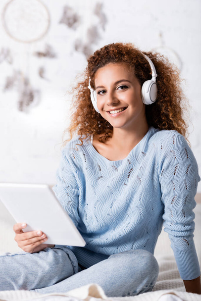Selective focus of curly woman in headphones looking at camera while holding digital tablet on bed  - Zdjęcie, obraz