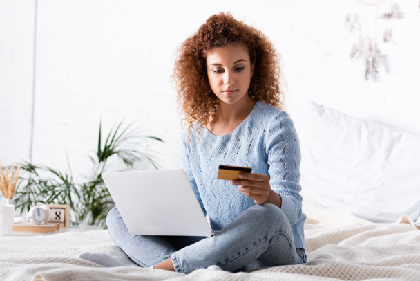 Selective focus of curly woman using laptop and credit card on bed  - Zdjęcie, obraz