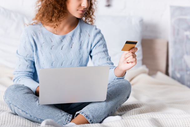 Vista recortada de la mujer joven usando el ordenador portátil y la tarjeta de crédito durante las compras en línea en el dormitorio  - Foto, imagen