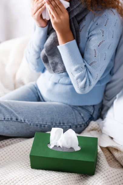 Selective focus of box with napkins on bed near sick woman in sweater and scarf  - Photo, Image