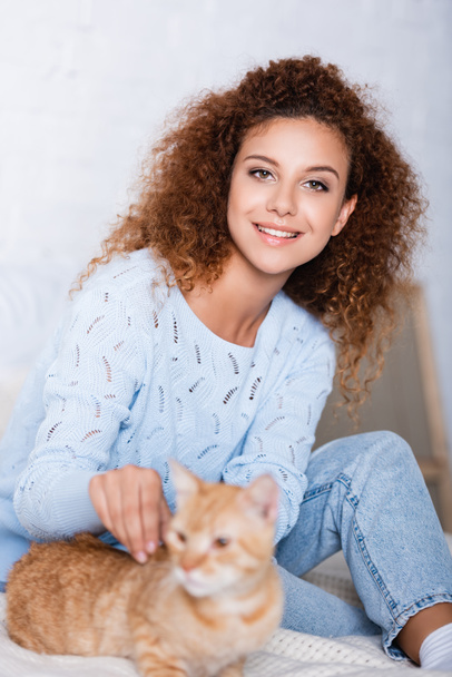 Selective focus of red haired woman in sweater looking at camera near cat at home  - Foto, Bild