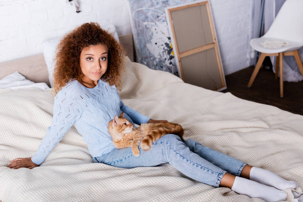 Red haired woman looking at camera near tabby cat on bed  - Photo, Image