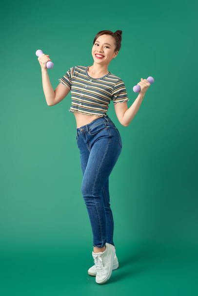 Attractive young Asian woman holding dumbell while standing over green background. Full length. - Φωτογραφία, εικόνα