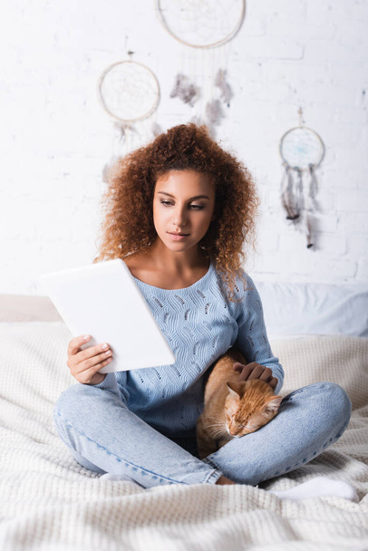Piros hajú nő pulóverben digitális tabletta és simogató macska az ágyban  - Fotó, kép