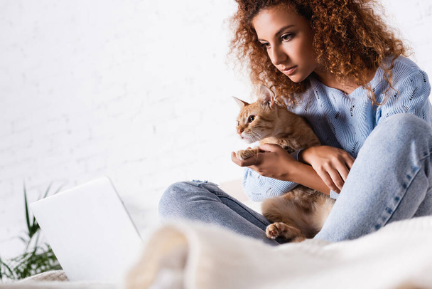 Selective focus of young red haired woman embracing cat near laptop at home  - Foto, imagen