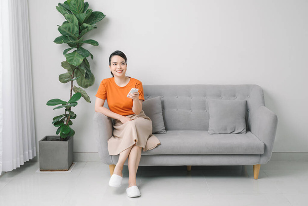 Junge glückliche Frau schaltet Klimaanlage ein, die auf Couch sitzt - Foto, Bild