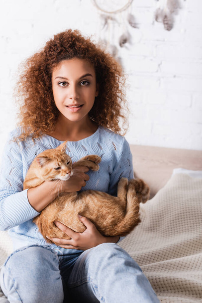 enfoque selectivo de la mujer joven mirando a la cámara mientras sostiene gato tabby en la cama  - Foto, Imagen