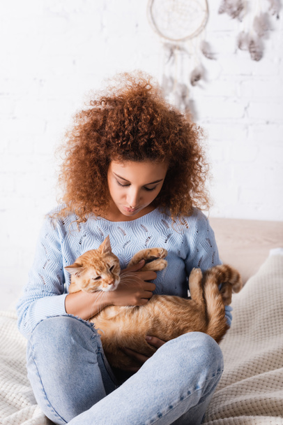 Selective focus of red haired woman holding tabby cat while sitting on bed  - Фото, изображение