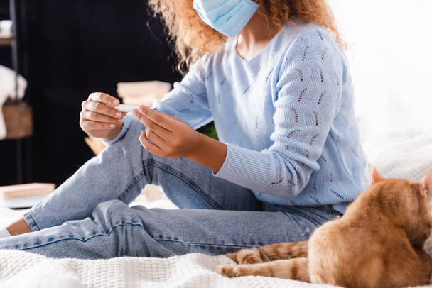 Selective focus of woman in medical mask holding thermometer near cat on bed  - Photo, Image