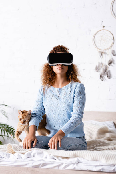 Selektiver Fokus der Frau im vr-Headset, die auf dem Bett neben gestromter Katze sitzt  - Foto, Bild