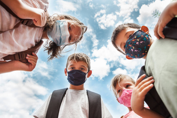 Vista de ángulo bajo de cuatro niños mirando hacia abajo a la cámara, usando máscaras protectoras de color. Están juntos en círculo con el cielo azul nublado detrás de ellos. - Foto, imagen