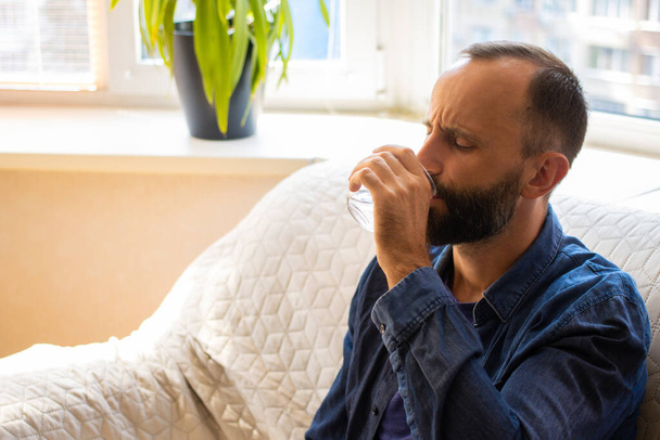 Millennial bearded man drinking glass of water, grimacing with tasteless medicine, taking antibiotic painkiller pill to relieve pain while sitting on couch at home. Emergency treatment concept. - Fotoğraf, Görsel