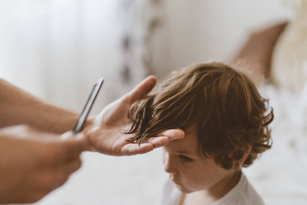 Father cuts her son hair in the room. Family during quarantine, Scissors and a comb in male hands - Valokuva, kuva