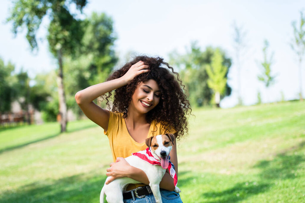 joyful woman touching curly hair while holding jack russell terrier dog in park - Photo, Image