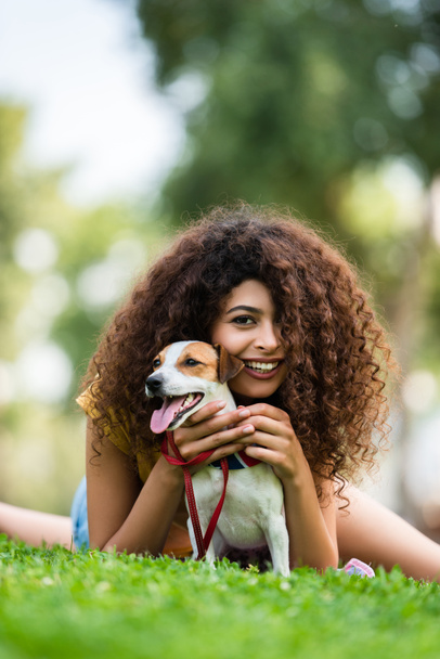 joyful curly woman looking at camera while relaxing with jack russell terrier dog on lawn - Foto, immagini
