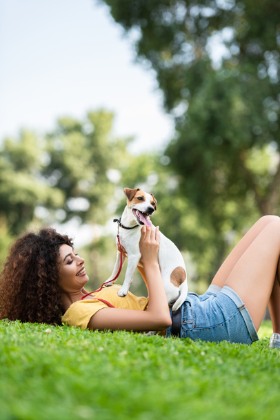 Yazlık kıyafetli neşeli kadın yeşil çimlerde Jack Russell Terrier köpeğiyle yatıyor. - Fotoğraf, Görsel