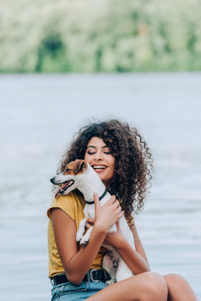 excited, curly woman in summer outfit embracing jack russell terrier dog near lake - Foto, Bild