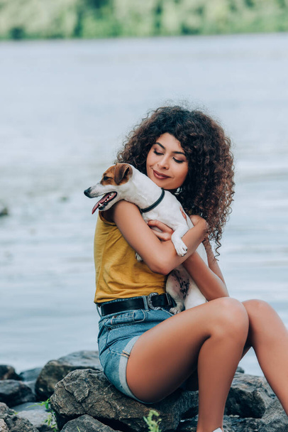 brunette, curly woman in summer outfit holding jack russell terrier dog while sitting on stones near river - Foto, Imagen