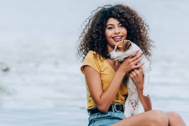 excited, curly woman in summer outfit embracing jack russell terrier dog near lake - Photo, Image