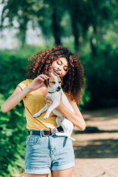 joyful woman in summer outfit stroking jack russell terrier dog in park - Fotó, kép