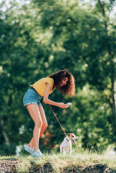 young woman in summer outfit laughing while taking photo of jack russell terrier dog in park - Fotó, kép