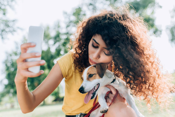 brunette, curly woman hugging jack russell terrier dog while taking selfie on mobile phone in park - Foto, Imagen