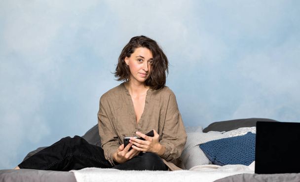 A beautiful brunette woman is lying on the bed in her bedroom with a smartphone in her hands. The girl is dressed in home clothes made of natural materials. The photo was taken from the front. - Foto, imagen
