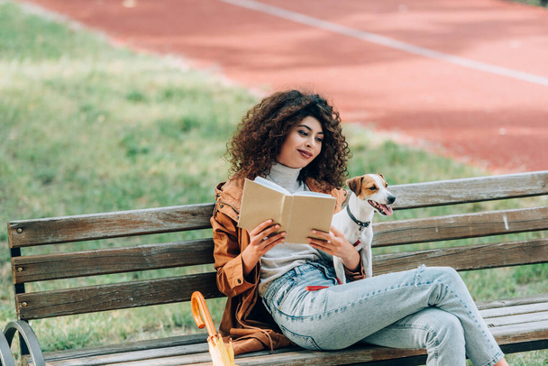stylish woman reading book while sitting on bench in park with jack russell terrier dog - Zdjęcie, obraz