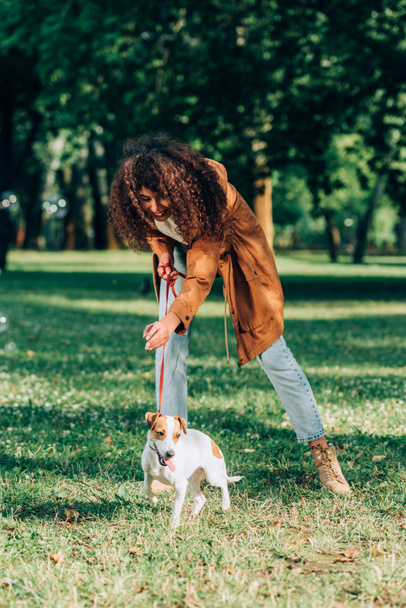 Yağmurluklu kıvırcık kadının parkta Jack Russell Terrier ile oynadığı seçici odak noktası.  - Fotoğraf, Görsel
