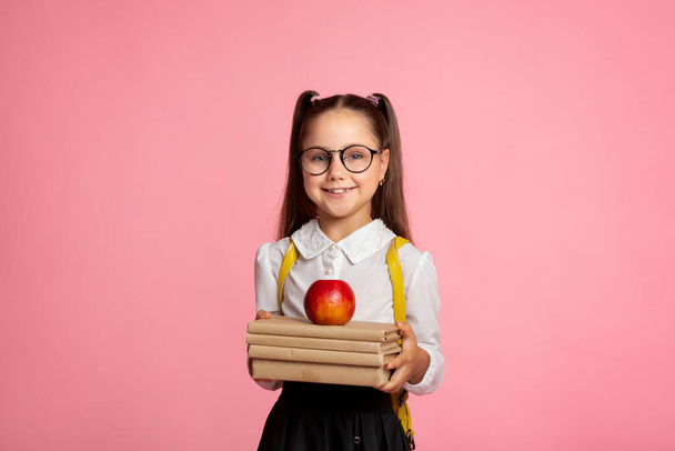 Early education concept. Smiling schoolgirl in glasses holds books and apple - Photo, Image