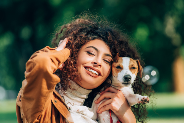 Selective focus of young curly woman with hand near head holding jack russell terrier outdoors  - Fotografie, Obrázek