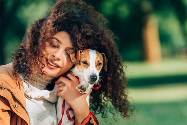 Junge Frau mit geschlossenen Augen umarmt Jack Russell Terrier im Freien  - Foto, Bild