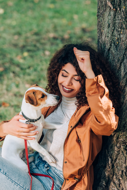 Curly woman in raincoat holding jack russell terrier near tree in park  - Foto, immagini