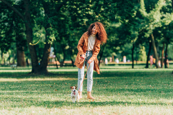 Selectieve focus van krullende vrouw in de herfst outfit houden riem tijdens een wandeling met jack russell terrier in het park  - Foto, afbeelding
