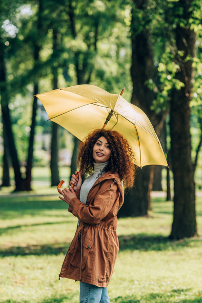 Σγουρή γυναίκα με αδιάβροχο κρατώντας ομπρέλα και κοιτάζοντας κάμερα στο πάρκο  - Φωτογραφία, εικόνα