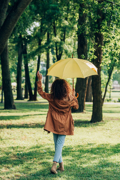 Back view of young woman in autumn outfit holding umbrella while walking in park  - Φωτογραφία, εικόνα