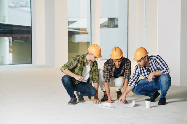 Serious young construction engineers in hardhats discussing construction plan on the floor in empty room - Photo, Image
