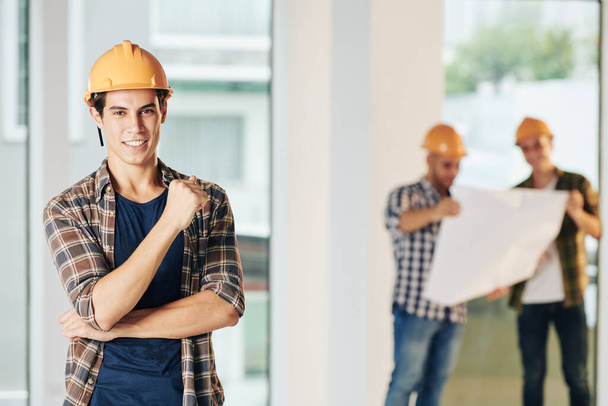 Portrét pohledného mladého stavebního inženýra se zubatým úsměvem stojícího na staveništi a hledícího do kamery - Fotografie, Obrázek