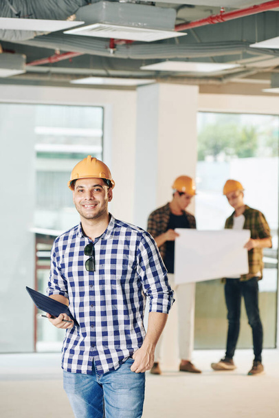 Jovem empreiteiro bonito positivo com tablet digital em pé dentro do edifício em construção - Foto, Imagem