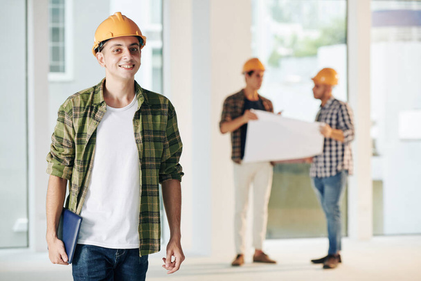 Young smiling middle eastern construction engineer in plaid shirt working on interior finishing with his team - Photo, Image