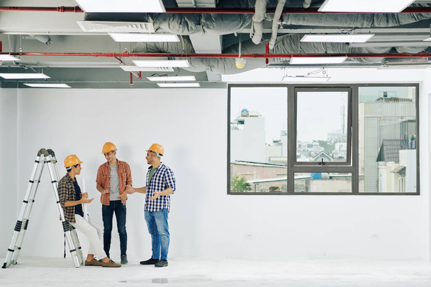 Jeunes ingénieurs de la construction en casquettes et chemises plaines discutant des nouvelles et des commérages pendant une courte pause au travail - Photo, image
