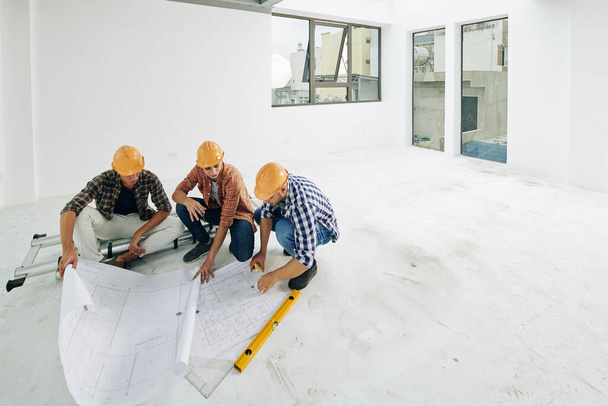 Команда молодых строителей в касках проверяет чертежи перед началом ремонта в квартире клиента - Фото, изображение
