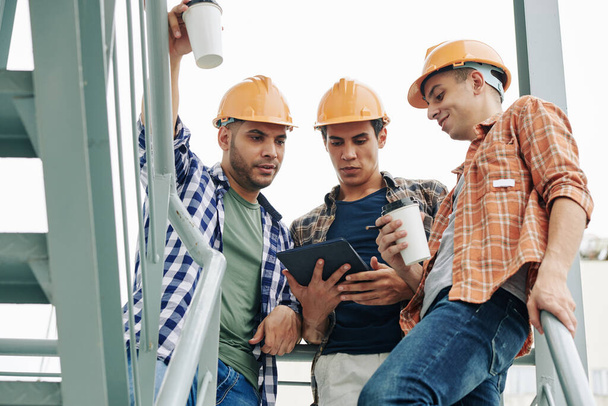 Young construction workers in hardhats drinking take away coffee and checking e-mail from contractor on tablet computer - Photo, Image