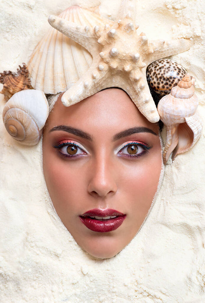 Beauty female face in sea sand and shells. Model with bright professional makeup posing beach. Summer travel beauty concept. Spa and wellness. Close up, selective focus. - 写真・画像