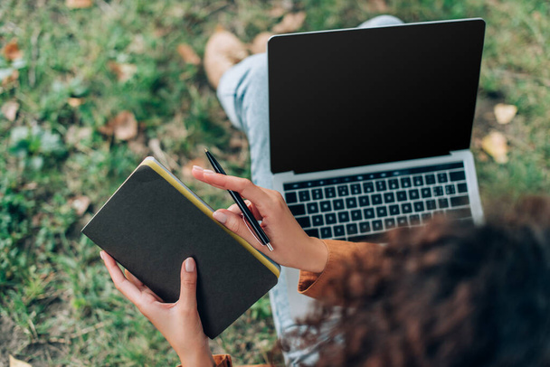 Widok kobiety trzymającej notatnik i długopis w pobliżu laptopa z pustym ekranem na trawie w parku  - Zdjęcie, obraz