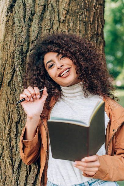 Selective focus of curly woman with notebook and pen looking at camera near tree  - Фото, зображення