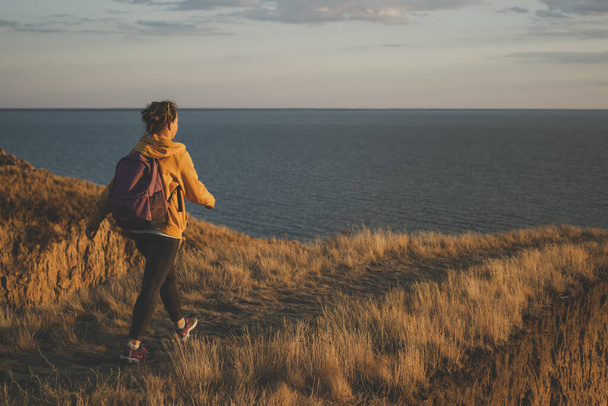 Widok z tyłu młodej kobiety podróżującej z plecakiem w żółtym kapturze spacerującej ścieżką szlaku na szczycie góry, wędrującej do klifu nad morzem podczas zachodu słońca. Miękki filtr. Koncepcja przygody i aktywnego stylu życia - Zdjęcie, obraz